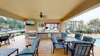 This outdoor amenity space is designed for relaxation and social gatherings, offering a thoughtfully arranged collection of features set against an attractive backdrop of multi-story brick and stone residential buildings. At the heart of the space is a resort-style swimming pool with a smooth paver surround, lined with cushioned lounge chairs and umbrella-shaded seating areas perfect for unwinding outdoors. Lush landscaping, including mature trees, manicured shrubs, and well-maintained green lawns, frames the pool deck and connecting courtyard pathways, creating a serene and inviting atmosphere. A spacious covered pavilion serves as the social centerpiece, equipped with a built-in outdoor fireplace, an outdoor kitchen and grill area, wall-mounted television, and a ceiling fan for year-round comfort. Plush striped lounge furniture and dining sets make this shaded retreat ideal for entertaining. Additional open-air courtyard areas feature decorative brick paving, café-style tables, and umbrellas nestled beneath mature trees. The surrounding multi-story buildings with balconies and bay windows provide an elegant architectural presence, while the carefully maintained grounds and water feature accents contribute to the polished, community-resort ambiance throughout the space.