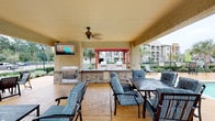 This outdoor amenity space is designed for relaxation and social gatherings, offering a thoughtfully arranged collection of features set against an attractive backdrop of multi-story brick and stone residential buildings. At the heart of the space is a resort-style swimming pool with a smooth paver surround, lined with cushioned lounge chairs and umbrella-shaded seating areas perfect for unwinding outdoors. Lush landscaping, including mature trees, manicured shrubs, and well-maintained green lawns, frames the pool deck and connecting courtyard pathways, creating a serene and inviting atmosphere. A spacious covered pavilion serves as the social centerpiece, equipped with a built-in outdoor fireplace, an outdoor kitchen and grill area, wall-mounted television, and a ceiling fan for year-round comfort. Plush striped lounge furniture and dining sets make this shaded retreat ideal for entertaining. Additional open-air courtyard areas feature decorative brick paving, café-style tables, and umbrellas nestled beneath mature trees. The surrounding multi-story buildings with balconies and bay windows provide an elegant architectural presence, while the carefully maintained grounds and water feature accents contribute to the polished, community-resort ambiance throughout the space.