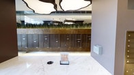 This well-appointed mailroom lobby combines functional design with refined aesthetics. The centerpiece of the space is a long wall of bronze-toned individual mailboxes, organized in a grid formation and set against a warm, metallic-finished panel that adds depth and sophistication. Above the mailboxes, a lush arrangement of ornamental grasses provides a striking natural accent, softening the otherwise structured and contemporary environment.

The flooring is finished in polished marble with a white and grey veining pattern, lending an upscale, clean feel to the space. A small informational display stand is positioned centrally on the floor, offering a practical focal point for residents. An outgoing mail slot is also integrated into the floor area, enhancing the room's utility.

The ceiling features sculptural, cloud-shaped pendant fixtures in a dark finish, adding an artistic and modern dimension to the overhead space. A large mirror on the back wall visually expands the room, reflecting the pendant lighting and greenery to create a sense of depth. Overall, the space balances practicality with a contemporary, upscale design sensibility.
