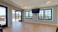 This single-floor rooftop space is designed as a versatile event and gathering venue, combining a bright, open interior with an accessible outdoor deck. The main interior area features warm hardwood-style flooring, neutral taupe walls, and a wall-mounted flat-screen display, making it well-suited for presentations, social gatherings, or private events. Large black-framed grid windows line the walls, flooding the interior with abundant natural light while offering open views of the surrounding landscape. A sliding glass door seamlessly connects the interior to the spacious outdoor deck, which is finished with composite decking and secured by modern metal railings. The deck provides an elevated outdoor experience with expansive open-sky views, comfortable seating, and access via both stairwell and elevator. A secondary area within the interior accommodates bar-height tables and stools, along with wall-mounted mini-split climate control units and elevator access, ensuring year-round comfort and convenience. Track lighting throughout the interior adds functional illumination, while the overall design strikes a balance between contemporary style and practical functionality. This rooftop space offers a polished, inviting environment ideal for hosting a wide range of events and gatherings.