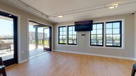 This single-floor rooftop space is designed as a versatile event and gathering venue, combining a bright, open interior with an accessible outdoor deck. The main interior area features warm hardwood-style flooring, neutral taupe walls, and a wall-mounted flat-screen display, making it well-suited for presentations, social gatherings, or private events. Large black-framed grid windows line the walls, flooding the interior with abundant natural light while offering open views of the surrounding landscape. A sliding glass door seamlessly connects the interior to the spacious outdoor deck, which is finished with composite decking and secured by modern metal railings. The deck provides an elevated outdoor experience with expansive open-sky views, comfortable seating, and access via both stairwell and elevator. A secondary area within the interior accommodates bar-height tables and stools, along with wall-mounted mini-split climate control units and elevator access, ensuring year-round comfort and convenience. Track lighting throughout the interior adds functional illumination, while the overall design strikes a balance between contemporary style and practical functionality. This rooftop space offers a polished, inviting environment ideal for hosting a wide range of events and gatherings.