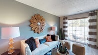 Located at 2645 Hard Rd in Columbus, Ohio, this well-appointed two-story home offers a comfortable and stylish living experience in the 43235 zip code. The living area immediately captures attention with its sage green accent wall, serving as a striking backdrop for a bold sunburst wooden mirror that adds warmth and artistic character to the room. A textured ceiling adds architectural depth overhead, while a large sliding glass door floods the space with natural light and provides a view of the outdoors, complemented by chic striped curtains that frame the window with casual elegance. The living room is thoughtfully arranged with a classic white sofa accented by vibrant orange, teal, and black throw pillows, creating a lively and inviting atmosphere. Touches of greenery, including potted topiary trees, bring a fresh, natural element indoors. The home spans two floors, offering generous living and private spaces that balance functionality with design. The overall ambiance throughout is warm, eclectic, and welcoming — a home that blends contemporary style with personal character in a prime Columbus location.