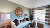 Located at 2645 Hard Rd in Columbus, Ohio, this well-appointed two-story home offers a comfortable and stylish living experience in the 43235 zip code. The living area immediately captures attention with its sage green accent wall, serving as a striking backdrop for a bold sunburst wooden mirror that adds warmth and artistic character to the room. A textured ceiling adds architectural depth overhead, while a large sliding glass door floods the space with natural light and provides a view of the outdoors, complemented by chic striped curtains that frame the window with casual elegance. The living room is thoughtfully arranged with a classic white sofa accented by vibrant orange, teal, and black throw pillows, creating a lively and inviting atmosphere. Touches of greenery, including potted topiary trees, bring a fresh, natural element indoors. The home spans two floors, offering generous living and private spaces that balance functionality with design. The overall ambiance throughout is warm, eclectic, and welcoming — a home that blends contemporary style with personal character in a prime Columbus location.