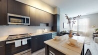 This single-floor residence showcases a cohesive contemporary design with light wood-look flooring and a neutral color palette that flows seamlessly throughout the space. The open-concept kitchen is a standout feature, equipped with full-height dark espresso cabinetry, light quartz countertops, a classic white subway tile backsplash, and a full suite of stainless steel appliances including a refrigerator, range, microwave, and dishwasher. A kitchen island provides additional prep and dining space, connecting naturally to the living area.

The living room is generously proportioned, benefiting from large windows dressed with sheer curtains that allow ample natural light to fill the space. The room comfortably accommodates a sectional sofa arrangement and a separate dining area, making it well-suited for both relaxing and entertaining.

The unit includes two bedrooms, each featuring windows with natural light and light wood-look flooring. The primary bedroom offers a calm, restful atmosphere with neutral tones and tasteful wall art. The second bedroom is compact yet functional.

The bathroom features a large glass-door walk-in shower with horizontal white tile surround, patterned floor tile, and a dark navy vanity with a white countertop, combining style with practicality.