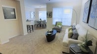 This single-floor residence offers an inviting, open-concept layout that effortlessly connects the main living areas. The living room is a comfortable and stylish centerpiece, featuring plush carpeting, a sleek oval coffee table, and a generously sized sofa accented with green and patterned throw pillows. A coordinating accent chair adds a pop of color and rounds out the seating arrangement. Large windows dressed with white horizontal blinds flood the space with abundant natural light, creating a warm and airy ambiance throughout. Crown molding along the ceiling adds a refined architectural touch. Flowing naturally from the living room, the dining area features an elegant round table surrounded by upholstered chairs, set beneath a modern pendant light fixture. A decorative mirror on the wall enhances the sense of depth and light in the dining space. Adjacent to the dining area, the kitchen counter with bar-height seating provides a casual spot for quick meals or entertaining guests. The neutral warm tones used throughout the walls and flooring create a cohesive, welcoming atmosphere that ties the entire living space together harmoniously.