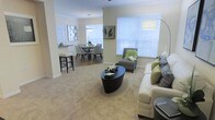 This single-floor residence offers an inviting, open-concept layout that effortlessly connects the main living areas. The living room is a comfortable and stylish centerpiece, featuring plush carpeting, a sleek oval coffee table, and a generously sized sofa accented with green and patterned throw pillows. A coordinating accent chair adds a pop of color and rounds out the seating arrangement. Large windows dressed with white horizontal blinds flood the space with abundant natural light, creating a warm and airy ambiance throughout. Crown molding along the ceiling adds a refined architectural touch. Flowing naturally from the living room, the dining area features an elegant round table surrounded by upholstered chairs, set beneath a modern pendant light fixture. A decorative mirror on the wall enhances the sense of depth and light in the dining space. Adjacent to the dining area, the kitchen counter with bar-height seating provides a casual spot for quick meals or entertaining guests. The neutral warm tones used throughout the walls and flooring create a cohesive, welcoming atmosphere that ties the entire living space together harmoniously.