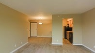 This single-floor space features an open-concept layout that connects the main living area to the kitchen through a half-wall partition, creating a sense of openness while maintaining a defined separation between the two zones. The living area is finished with wall-to-wall carpeting in a neutral tone, contributing to a warm and comfortable atmosphere. Warm beige and sage green walls complement the space throughout, lending a calm, cohesive aesthetic.

A pendant light fixture hangs from the ceiling near the entryway, providing focused ambient lighting and adding a subtle decorative element. The front door features a classic paneled design framed by dark trim, offering a clean and traditional look at the entry point.

The kitchen is partially visible through the opening and is finished with tile flooring, medium-toned wooden cabinetry, and black appliances including a refrigerator and a range. The kitchen benefits from its own overhead lighting, creating a warm, inviting cooking space. White baseboards run throughout, offering a clean contrast against the wall colors. Overall, the space presents a functional and well-organized single-level layout suitable for everyday living.