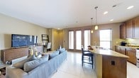 This single-floor space showcases an open-concept layout that integrates the living and kitchen areas into one cohesive, functional environment. The living area is anchored by a plush gray sectional sofa and accented by a lean-style media shelf unit, creating a casual yet polished atmosphere. Glossy large-format white tile flooring runs throughout, reflecting natural light and enhancing the sense of openness.

The kitchen features a granite-topped peninsula with dark cabinetry and seating for two on sleek bar stools, making it well-suited for casual dining or entertaining. Pendant lighting with warm amber glass shades hangs above the counter, adding a refined focal point. A stainless steel undermount sink and modern faucet complete the functional kitchen details.

Natural light is a defining characteristic of this space, flowing in generously through a set of French doors and flanking windows. The French doors open outward to an exterior view, offering a seamless connection to the outdoors. Neutral warm-toned walls complement the light flooring and dark cabinetry, resulting in a balanced, contemporary aesthetic throughout.