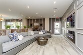 This single-floor amenity space is thoughtfully designed to serve as a dynamic social hub, blending comfort and functionality across several distinct zones. The entrance lobby impresses immediately with floor-to-ceiling glass walls, light wood-look flooring, and a dedicated mailroom, creating a clean and welcoming first impression. Beyond the entry, a spacious lounge area features a large sectional sofa, a round ottoman coffee table, and accent walls with layered textures and artwork, offering a relaxed retreat for residents. Adjacent to the lounge, a fully equipped community kitchen showcases warm wood cabinetry, stone countertops, stainless steel appliances including a dishwasher and microwave, open black-metal shelving, and pendant lighting — ideal for entertaining or casual use. A generously sized bar counter with stone surfaces and bar-height seating serves as a central social gathering point. The dining area provides multiple seating configurations, including banquette-style booth seating and standard table-and-chair arrangements beneath recessed lighting. A game room section features a full-size billiards table, adding a recreational element to the space. Throughout, large-format tile and wood-look flooring unify the open layout, while wall-mounted televisions, curated artwork, and lush greenery contribute to the space's polished, contemporary character.