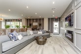 This single-floor amenity space is thoughtfully designed to serve as a dynamic social hub, blending comfort and functionality across several distinct zones. The entrance lobby impresses immediately with floor-to-ceiling glass walls, light wood-look flooring, and a dedicated mailroom, creating a clean and welcoming first impression. Beyond the entry, a spacious lounge area features a large sectional sofa, a round ottoman coffee table, and accent walls with layered textures and artwork, offering a relaxed retreat for residents. Adjacent to the lounge, a fully equipped community kitchen showcases warm wood cabinetry, stone countertops, stainless steel appliances including a dishwasher and microwave, open black-metal shelving, and pendant lighting — ideal for entertaining or casual use. A generously sized bar counter with stone surfaces and bar-height seating serves as a central social gathering point. The dining area provides multiple seating configurations, including banquette-style booth seating and standard table-and-chair arrangements beneath recessed lighting. A game room section features a full-size billiards table, adding a recreational element to the space. Throughout, large-format tile and wood-look flooring unify the open layout, while wall-mounted televisions, curated artwork, and lush greenery contribute to the space's polished, contemporary character.