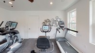 Located at 5313 Fairfield Rd in Columbia, SC, this dedicated fitness center offers a well-equipped, single-floor workout facility designed to support a well-rounded exercise routine. The space is finished with durable rubber-flecked flooring throughout, providing a safe and functional surface for a variety of activities.

The cardio area is stocked with a quality selection of True-brand equipment, including treadmills, an elliptical trainer, and a stationary recumbent bike, each outfitted with interactive touchscreen displays for an engaging workout experience. A ceiling fan overhead aids in air circulation during intense sessions. A decorative circular mirror wall accent adds a stylish touch to the otherwise clean, minimalist interior.

A separate strength-training area houses a tiered dumbbell rack with a comprehensive range of free weights, as well as an adjustable workout bench, offering ample options for resistance training. Recessed lighting and a ceiling fan keep the space bright and comfortable.

Large double-hung windows line the walls, inviting abundant natural light and providing views of the surrounding neighborhood. A glass-paneled exterior door with an automatic opener allows for easy access directly from outside. The crisp white walls and clean layout create an open, motivating atmosphere well-suited for daily fitness use.