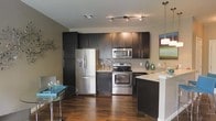 This single-floor space showcases a thoughtfully designed open-concept layout where the kitchen and dining area share a seamless connection. The kitchen is a standout feature, outfitted with sleek, dark espresso cabinetry that extends to the ceiling, providing generous storage. Stainless steel appliances, including a French door refrigerator with an ice and water dispenser, a built-in microwave, and a gas range, lend the space a polished, contemporary look. The light granite countertops offer a striking contrast against the dark cabinetry, and a mosaic tile backsplash adds a subtle decorative touch. A peninsula with bar-height seating extends the counter space and doubles as a casual dining option. Track lighting mounted on the ceiling, complemented by a pendant light over the peninsula, ensures the kitchen is well-illuminated. Rich, dark hardwood flooring runs throughout, anchoring the space with warmth and depth. The dining area accommodates a glass-top table with seating, creating an intimate setting for meals. The overall ambiance is modern and sophisticated, balancing dark and light tones for a visually dynamic interior.