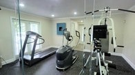 This single-floor fitness room is thoughtfully designed to serve as a private workout space. The room features rubber flooring, which provides durability and cushioning underfoot, making it well-suited for high-impact exercise. Recessed lighting in the white ceiling, accented by crown molding, keeps the space bright and well-illuminated. Large windows along the wall allow natural light to filter in, contributing to an open and energizing atmosphere. Full-length mirrors line one wall, visually expanding the room while allowing users to monitor their form during workouts. The space is equipped with cardio machines, including a treadmill and an elliptical stepper, positioned to make efficient use of the layout. A mounted screen and camera tripod suggest the room may also be used for virtual training or workout streaming. Two interior doors offer convenient access points, and the neutral wall tones paired with a single teal accent panel add a subtle pop of color. Overall, the room strikes a balance between functional fitness infrastructure and a clean, comfortable aesthetic.