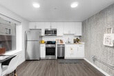 This contemporary single-floor unit is thoughtfully designed with modern finishes and functional living spaces. The kitchen serves as a standout feature, outfitted with a full suite of stainless steel appliances — including a refrigerator, gas range, built-in microwave, and dishwasher — complemented by sleek white cabinetry, light countertops, and a textured accent wall that adds visual depth. Rich wood-look flooring runs throughout the unit, creating a cohesive and warm aesthetic. The bedroom is a cozy, well-defined space featuring a window that allows in natural light and offers a view of the surrounding neighborhood. The neutral tones of the walls are accented by the same textured wall treatment seen in the kitchen, maintaining a consistent design language. A dedicated area within the unit houses an in-unit washer/dryer combination, adding everyday convenience, while large windows in the living area bring in ample natural light and views of the exterior. The bathroom is clean and functional, featuring a full-size soaking tub with a shower and curtain rod, finished in crisp white tones. Overall, the unit presents a modern, practical, and well-appointed living space suitable for comfortable day-to-day living.