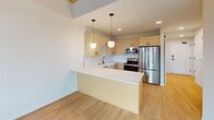This 560-square-foot, one-bedroom, one-bathroom apartment in Madison, WI (53703) delivers a modern, well-appointed living experience across a single floor. The kitchen is a standout feature, showcasing light maple cabinetry with matte black hardware, a classic white farmhouse sink with a black matte faucet, a white quartz peninsula with pendant globe lighting, and a full suite of stainless steel appliances including a range, microwave, and refrigerator. Light hardwood flooring flows throughout the main living and kitchen areas, enhancing the bright, open feel. The 130-square-foot primary bedroom features plush carpet, crisp white walls, and a large floor-to-ceiling window framed by an exposed wood beam, offering sweeping city views. A dedicated closet provides organized storage with a built-in shelving system featuring metal wire shelves, drawers, and hanging rods. The bathroom continues the modern aesthetic with a wood-framed double vanity, arched mirror with integrated sconce lighting, white quartz countertop, matte black fixtures, and a glass-enclosed shower. A hallway connects the rooms efficiently, completing this sleek and functional urban residence.