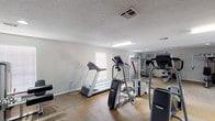 This single-floor fitness center on Parkmount Boulevard in Baton Rouge, Louisiana, offers a well-equipped space designed to support a comprehensive workout experience. The main exercise area is generously sized, providing ample room to move freely between a variety of cardio and strength-training machines. Equipment includes multiple treadmills, elliptical trainers, a stationary bike, and a Precor cross-trainer, alongside a multi-function cable and weight machine, a rowing-style resistance machine, an adjustable weight bench, and a dumbbell rack. The flooring throughout the workout area combines durable hardwood-style sections with modular carpet tiles, offering both stability and comfort underfoot. Natural light filters in through several windows fitted with blinds, supplemented by overhead fluorescent and recessed lighting to keep the space well-illuminated at all times. A textured ceiling adds a classic finish to the interior. Adjacent to the main workout floor is a separate lounge or media room, featuring wall-to-wall carpet tile flooring, a large wall-mounted flat-screen television, a water cooler, and seating. A set of glass-paned double doors at the rear of the fitness area opens directly to an outdoor pool area, creating a seamless connection between indoor fitness and outdoor recreation.