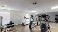 This single-floor fitness center on Parkmount Boulevard in Baton Rouge, Louisiana, offers a well-equipped space designed to support a comprehensive workout experience. The main exercise area is generously sized, providing ample room to move freely between a variety of cardio and strength-training machines. Equipment includes multiple treadmills, elliptical trainers, a stationary bike, and a Precor cross-trainer, alongside a multi-function cable and weight machine, a rowing-style resistance machine, an adjustable weight bench, and a dumbbell rack. The flooring throughout the workout area combines durable hardwood-style sections with modular carpet tiles, offering both stability and comfort underfoot. Natural light filters in through several windows fitted with blinds, supplemented by overhead fluorescent and recessed lighting to keep the space well-illuminated at all times. A textured ceiling adds a classic finish to the interior. Adjacent to the main workout floor is a separate lounge or media room, featuring wall-to-wall carpet tile flooring, a large wall-mounted flat-screen television, a water cooler, and seating. A set of glass-paned double doors at the rear of the fitness area opens directly to an outdoor pool area, creating a seamless connection between indoor fitness and outdoor recreation.