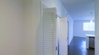 This single-floor space presents a clean, well-maintained interior defined by its minimalist aesthetic and functional design. The corridor area is immediately notable for its crisp white walls and smooth ceiling, which together create a bright and airy atmosphere throughout. White louvered bifold doors line the hallway, offering a classic yet practical design element that adds subtle visual texture while maintaining a cohesive, uniform look. The louvered panels allow for ventilation while neatly concealing storage or utility areas behind them. The flooring along the corridor features a muted, dark-toned surface that provides a grounding contrast to the predominantly white walls and doors, contributing to a balanced visual composition. Ceiling-mounted vents are integrated discreetly into the design, reflecting attention to detail in the overall layout. The hallway stretches with a sense of orderly depth, suggesting a thoughtfully planned floor plan. The overall ambiance of the space is clean, functional, and understated, making it well-suited for a variety of uses. The consistent use of white throughout the interior enhances the sense of openness and keeps the environment feeling light and organized.