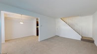 This two-floor residence offers a clean, functional layout designed for comfortable everyday living. The main level centers around a generously sized living area finished with neutral wall-to-wall carpeting that lends warmth underfoot. Bright white walls reflect light throughout the space, contributing to an open and airy atmosphere. A classic pendant light fixture anchors one corner of the room, providing both task and ambient illumination. An open doorway connects the living area seamlessly to the kitchen, which features dark cabinetry and direct access to additional living spaces. A closet door on the main level provides convenient storage. The staircase, fitted with a gold-toned banister railing, rises gracefully from the living area to the upper floor, offering a traditional architectural detail that adds visual interest to the space. The overall design is straightforward and well-maintained, with a neutral palette throughout that provides a versatile backdrop for a variety of interior styles. The two-floor layout offers a natural separation between living and private spaces, making this a practical and welcoming home.