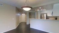 This single-floor residence offers an open-concept layout that connects the living area, kitchen, and entryway in a cohesive, functional design. Dark hardwood floors run throughout, providing a striking contrast against the neutral beige walls and white trim work.

A prominent bowl-style pendant light fixture hangs from the textured ceiling, serving as a focal point above the main living area, while recessed lighting and track lighting illuminate the kitchen space. The kitchen features white cabinetry, a stainless steel refrigerator, and a raised breakfast bar with a light-colored countertop that doubles as a partition between the kitchen and living areas.

Toward the rear of the unit, a dedicated in-unit laundry area is neatly tucked behind the kitchen, offering added convenience. The entryway is framed by a classic paneled door with transom windows above, allowing natural light to filter into the space. A secondary door along the hallway provides additional access or storage options. Overall, the space maintains a clean, neutral palette with practical finishes throughout.