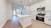 This 570-square-foot, one-bedroom, one-bathroom apartment in North Augusta, SC delivers a clean, modern aesthetic across a single-floor layout. The kitchen serves as the centerpiece of the unit, featuring white shaker cabinets, a white subway tile backsplash, stainless steel appliances including a range and French door refrigerator, and a large central island with a white quartz countertop and an undermount sink with a matte black faucet. Pendant lighting above the island and recessed ceiling lights provide ample illumination. Light wood-look flooring flows consistently throughout the entire unit, creating a cohesive and airy atmosphere. The kitchen opens toward a living area with access to a glass-panel exterior door, allowing natural light to fill the space. The 130-square-foot bedroom offers a bright, neutral environment with dual windows fitted with blinds. The bathroom is well-appointed with a white vanity, quartz countertop, matte black hardware, a framed mirror, and a glass-enclosed walk-in shower with tile surround. A dedicated closet provides additional storage. The unit's consistent white walls, clean lines, and contemporary hardware selections give the entire space a fresh, move-in-ready feel.
