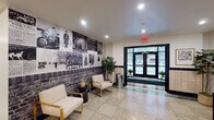 This building's ground-floor common areas masterfully blend historic architectural character with contemporary updates. The main lobby serves as a welcoming focal point, featuring polished terrazzo floors in a checkered pattern and original large-format wall tiles accented by dark contrasting borders — details that speak to the building's storied past. A striking black-and-white photographic mural spanning one wall celebrates the building's history, complemented by tasteful seating arrangements with modern gold-framed chairs and a console table.

The entrance vestibule is framed by classic black-trimmed glass double doors that invite ample natural light from the street. Adjacent to the lobby, a secondary entry area features warm wood-tone flooring, black mailbox banks, parcel lockers, and a glass-partitioned office space, providing practical amenities in a clean, modern aesthetic.

Connecting corridors extend the building's classic character through original glazed yellow brick walls and terrazzo flooring, lending a sense of institutional heritage throughout. A coffee station with a rustic wooden sideboard adds a welcoming, community-oriented touch near corridor access points. Together, these spaces create a cohesive environment that honors architectural tradition while meeting contemporary functional needs.