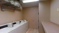 This single-floor laundry room is a functional, no-frills utility space designed to accommodate everyday washing and drying needs. The room features multiple side-by-side washing machines and dryers lined along one wall, providing ample capacity for regular laundry tasks. A built-in folding counter runs along the opposite wall, offering a convenient surface for sorting and folding clothes. Overhead, fluorescent lighting illuminates the entire space evenly, ensuring good visibility throughout. A metal shelf bracket system is mounted above the machines, providing additional storage for laundry supplies. The walls are finished in a neutral beige tone, and the floor is covered in light-colored ceramic tile, making the space easy to maintain and clean. A solid door with a secure lock provides controlled access to the room, suggesting this may be a shared laundry facility. A small utility panel or intercom device is mounted on the wall near the door, likely for building access or operational controls. The overall layout is practical and straightforward, maximizing efficiency within a modest footprint.