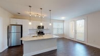 This well-appointed single-floor residence showcases a thoughtfully designed open-concept layout that seamlessly connects the kitchen, dining, and living areas. The kitchen is a standout feature, outfitted with crisp white shaker-style cabinetry, granite countertops, a white penny-tile backsplash, and a full suite of stainless steel appliances including a refrigerator, electric range, and built-in microwave. A central island with pendant lighting and an integrated sink serves as both a functional workspace and a natural gathering point. Rich, dark hardwood-style flooring runs throughout the main living areas, complemented by neutral walls and an abundance of natural light streaming through multiple large windows fitted with horizontal blinds. An arched entryway adds an elegant architectural detail to the hallway, which leads to the bedrooms. The bedrooms feature plush carpeting, ceiling fans, and natural light from well-placed windows. Two bathrooms are finished with light tile flooring and granite-topped vanities; one bathroom includes a tub-shower combination with clean white tile surround, while the other features a dark-toned vanity cabinet with matching wall-mounted storage. The overall aesthetic is clean, contemporary, and move-in ready.