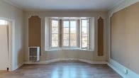 This single-floor space showcases a blend of classic architectural character and functional design. Throughout the living areas, warm beige walls are accentuated by elegant white decorative moldings and panel frames, lending a timeless, period-appropriate aesthetic. Light hardwood-style flooring runs consistently across the rooms, enhancing the sense of openness and cohesion.

A standout feature of the main living space is a generous bay window that curves outward, flooding the room with natural light and offering views of tree-lined surroundings. Ceiling fans are present in the living areas, contributing to both comfort and airflow. Crown molding adds a refined finishing touch throughout.

The bedroom area is notable for its built-in wardrobe closets framed by white paneled doors with mirrored accents, providing ample storage. An adjacent vanity area features a white dresser with a wall-mounted mirror, offering a dedicated grooming space.

The bathroom retains a distinctive retro character, highlighted by mint green ceramic wall tiles trimmed with black accent borders. A recessed medicine cabinet with a mirrored door, a pedestal-style sink, and a window providing natural ventilation complete this vintage-inspired space.

The overall layout, as revealed by the dollhouse view, is well-organized and flows naturally from room to room, making efficient use of the available space.