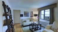 This single-floor space showcases a thoughtfully designed living area that balances comfort and style. The room features warm hardwood flooring throughout, providing a natural foundation that complements the neutral color palette of cream and taupe tones on the walls and furnishings. A light sage-green sofa serves as the focal point of the seating area, accented with striped throw pillows and flanked by two matching black floor lamps that offer both ambient and task lighting. A textured area rug grounds the seating arrangement, which includes a pair of dark espresso-finished nesting tables topped with decorative candles and a fresh floral arrangement. Two framed abstract artworks hang above the sofa, adding visual interest and a touch of color to the otherwise understated walls. Natural light filters into the space through a double-hung window dressed with neutral drapery panels, offering a glimpse of greenery outside. A white door with classic trim provides access to an adjoining room. A radiator beneath the window suggests traditional building infrastructure, while the overall composition of the space reflects a curated, elegant aesthetic.