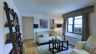 This single-floor space showcases a thoughtfully designed living area that balances comfort and style. The room features warm hardwood flooring throughout, providing a natural foundation that complements the neutral color palette of cream and taupe tones on the walls and furnishings. A light sage-green sofa serves as the focal point of the seating area, accented with striped throw pillows and flanked by two matching black floor lamps that offer both ambient and task lighting. A textured area rug grounds the seating arrangement, which includes a pair of dark espresso-finished nesting tables topped with decorative candles and a fresh floral arrangement. Two framed abstract artworks hang above the sofa, adding visual interest and a touch of color to the otherwise understated walls. Natural light filters into the space through a double-hung window dressed with neutral drapery panels, offering a glimpse of greenery outside. A white door with classic trim provides access to an adjoining room. A radiator beneath the window suggests traditional building infrastructure, while the overall composition of the space reflects a curated, elegant aesthetic.