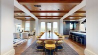 Located at 8525 Connecticut Ave in Chevy Chase, Maryland, this refined single-floor space exemplifies upscale design through a seamless blend of classic and contemporary elements. Rich wood coffered ceilings with recessed lighting are a defining architectural feature throughout, lending warmth and a sense of grandeur to every area.

The main gathering area centers on a substantial wood conference table surrounded by gold-framed chairs upholstered in soft neutral tones, ideal for formal meetings or group events. Floor-to-ceiling arched windows and glass doors flood the space with natural light while offering views of the surrounding landscape and an adjoining outdoor terrace.

A separate lounge area provides a more relaxed setting, featuring plush seating, wood-paneled walls, and direct terrace access via draped glass doors. A secondary dining nook with a round table and woven chairs adds versatility to the layout.

The fully equipped kitchen showcases deep-toned cabinetry with fluted detailing, a white stone-topped island with bar seating, and a statement multi-pendant brass-and-black light fixture. Wide-plank hardwood flooring flows throughout, unifying the space. A bright, art-accented corridor connects all areas, completing this polished and functional environment.