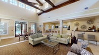 This well-appointed single-floor community clubhouse offers an inviting and polished atmosphere designed for comfort and socializing. The centerpiece of the space is the grand main lounge, where soaring ceilings adorned with striking exposed dark wood beams command attention. Tall clerestory windows flood the area with natural light, enhancing the open, airy feel. Plush sage green sofas and a glass coffee table are arranged atop a patterned area rug, creating a warm and welcoming seating area. Adjacent to the main lounge, a reception and lobby area features additional seating with accent chairs and decorative wall art, anchored by a staffed front desk. A dedicated bar area includes a long granite-topped counter with bar stool seating, pendant lighting, and multiple wall-mounted televisions, making it ideal for casual gatherings. A cozy fireplace nook showcases a built-in log storage alcove and tufted ottoman seating beneath framed artwork. The dining room offers a polished oval table with upholstered chairs and direct access to an outdoor pool area through elegant glass-paneled doors, framed by large windows with views of lush greenery. Throughout, warm neutral tones, crown molding, and carefully curated décor contribute to a refined yet relaxed ambiance.