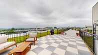 Located at 1818 N 53rd St in Philadelphia, Pennsylvania, this rooftop terrace offers an expansive outdoor amenity space with a striking combination of design and functionality. The deck features a bold checkered tile pattern in gray and white, extending the length of the rooftop and providing a generous area for outdoor gatherings and relaxation.

A standout feature of this rooftop is its living green roof, which spans a substantial portion of the space and is bordered by sleek black metal railings. The lush, low-maintenance sedum greenery adds a vibrant, eco-conscious element to the rooftop's overall design. Patio heaters are positioned throughout the space, extending its usability across multiple seasons.

Seating is thoughtfully arranged along the terrace, including wooden lounge chairs, benches, and dining tables, creating distinct zones for lounging and socializing. Potted plants accent the space with additional greenery.

Panoramic views are a defining characteristic of this rooftop, with the Philadelphia city skyline visible in the distance and wide, open sightlines across the surrounding neighborhood. The space is enclosed by sturdy black metal railings, ensuring both safety and unobstructed views. Access is provided via an exterior door, and the overall layout offers a well-organized, inviting outdoor retreat above the city.