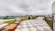 Located at 1818 N 53rd St in Philadelphia, Pennsylvania, this rooftop terrace offers an expansive outdoor amenity space with a striking combination of design and functionality. The deck features a bold checkered tile pattern in gray and white, extending the length of the rooftop and providing a generous area for outdoor gatherings and relaxation.

A standout feature of this rooftop is its living green roof, which spans a substantial portion of the space and is bordered by sleek black metal railings. The lush, low-maintenance sedum greenery adds a vibrant, eco-conscious element to the rooftop's overall design. Patio heaters are positioned throughout the space, extending its usability across multiple seasons.

Seating is thoughtfully arranged along the terrace, including wooden lounge chairs, benches, and dining tables, creating distinct zones for lounging and socializing. Potted plants accent the space with additional greenery.

Panoramic views are a defining characteristic of this rooftop, with the Philadelphia city skyline visible in the distance and wide, open sightlines across the surrounding neighborhood. The space is enclosed by sturdy black metal railings, ensuring both safety and unobstructed views. Access is provided via an exterior door, and the overall layout offers a well-organized, inviting outdoor retreat above the city.