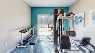 Located at 1045 Floribunda Way in Webster, NY, this single-floor fitness center offers a well-appointed workout space designed to support a range of exercise routines. The entry corridor makes a striking first impression with bold, geometric-patterned wallpaper in deep navy and gold tones, transitioning seamlessly into the main workout area via a classic white paneled door. The fitness floor features a stylish patterned carpet in teal and grey tones that lends the space a modern, energetic feel. Walls are finished in a warm neutral tone, with one accent wall painted a vibrant teal that adds visual energy to the room. The space is equipped with a variety of cardio machines, including a treadmill and stationary bikes, as well as a multi-function Body-Solid strength training station and a full dumbbell rack with a flat bench. A stability ball and wall-mounted TV round out the amenities. Natural light fills the room through multiple windows and a sliding glass door that opens to views of an outdoor pool and surrounding greenery. Fitness-themed wall art adds a finishing decorative touch. The drop ceiling with recessed panel tiles provides a clean, finished aesthetic throughout this practical and inviting community fitness facility.
