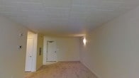 This single-floor space presents a straightforward and functional layout with a classic drop tile ceiling that gives the interior a clean, structured appearance. The soft carpeted flooring in neutral tones adds a layer of comfort underfoot and contributes to the room's quiet, understated ambiance. Warm-toned walls complement the overall palette, creating a simple yet cohesive interior environment. A wall-mounted sconce provides supplemental lighting, casting a soft glow across the space. The room features a standard interior door, along with what appears to be a wall-mounted panel, suggesting practical utility provisions built into the design. The space is open and unobstructed, lending itself well to a variety of uses and configurations. While modest in its finishes, the layout is efficient and well-organized, making it a versatile environment suitable for a range of functions. The drop ceiling, neutral carpeting, and wall lighting together create a calm, contained atmosphere that prioritizes function and ease of use.