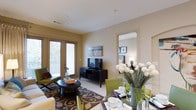 This single-floor residence in Dallas offers a thoughtfully designed layout that seamlessly blends comfort and functionality. The open-concept living and dining area serves as the heart of the home, bathed in natural light that filters through French doors leading to a private outdoor patio. The living space features warm neutral wall tones and light-colored flooring, creating a bright and inviting atmosphere throughout. A decorative area rug anchors the seating arrangement, which centers around a round coffee table, lending the space a cozy yet polished feel. The dining area is positioned adjacent to the living room, allowing for a natural flow between the two spaces, and is furnished with a glass-topped table surrounded by green upholstered chairs — a stylish focal point that adds a pop of color to the neutral palette. A mirror mounted near the bedroom entry enhances the sense of depth and openness. The bedroom, visible through a wide doorway, continues the refined aesthetic with coordinated furnishings and soft lighting. Throughout the residence, clean lines, tasteful décor, and an abundance of natural light combine to create a space that feels both elegant and comfortable — ideal for everyday living in the heart of Dallas.