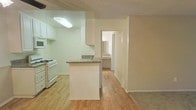 This single-floor residence offers a functional and straightforward layout designed for comfortable everyday living. The kitchen is equipped with white cabinetry, a gas range, a built-in microwave, and a peninsula with a granite-style countertop that provides additional prep space and a natural divider between the kitchen and the adjacent living area. A textured ceiling adds a classic touch overhead, while recessed flush lighting keeps the kitchen bright and well-illuminated. Light hardwood flooring extends throughout the kitchen and hallway, creating a cohesive and easy-to-maintain surface underfoot. A central hallway leads to the remaining rooms, including what appears to be a bathroom and additional living spaces visible in the background. The neutral, light-toned walls throughout contribute to an open and airy feel, maximizing the sense of space within the home. The transition from hardwood flooring to carpet hints at a designated living or bedroom area just beyond the main corridor. Overall, the space presents a clean, practical interior well-suited for those seeking a no-frills, move-in-ready home environment.