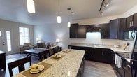 This well-appointed single-floor space showcases a thoughtfully designed open-concept layout that integrates the kitchen, dining, and living areas into one cohesive environment. The kitchen is a standout feature, boasting rich dark cabinetry that contrasts beautifully with light speckled granite countertops. A generous kitchen island serves as both a prep space and casual dining surface, complete with place settings and pendant lighting overhead. A built-in wine rack adds a sophisticated touch to the upper cabinetry, while a double sink and dishwasher offer practical functionality. Track lighting along the ceiling provides ample illumination throughout the kitchen zone. The open dining area transitions naturally into the living room, which is furnished with a comfortable neutral-toned sofa and coordinating accent chairs arranged around a dark wood coffee table. Warm table lamps contribute to the inviting ambiance. Hardwood-style flooring runs throughout, tying the spaces together with a clean, modern aesthetic. The overall design balances style and functionality, making this space ideal for both everyday living and entertaining.