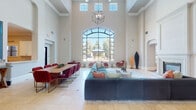 This single-floor space makes an immediate impression through its grand entryway corridor, lined with floor-to-ceiling French doors and glass panels on both sides, topped by a striking arched transom window. Natural light pours in across the slate-tiled floor, setting a refined tone from the moment of entry.

The main gathering area commands attention with dramatically high ceilings adorned with crown molding and clerestory windows that flood the room with daylight. A large arched window wall frames views of a lush outdoor courtyard, bringing the exterior landscape into the interior experience. Suspended from above, a geometric cube-style chandelier serves as a sculptural focal point.

The lounge zone centers around a generously sized sectional sofa in deep teal velvet, paired with accent chairs in warm tones and a marble-top coffee table. A stately white fireplace with arched detailing anchors one wall, further enhancing the space's elegance.

Adjacent to the lounge, a long communal dining table surrounded by rich burgundy velvet chairs accommodates group gatherings. A vibrant large-scale abstract artwork and a pass-through kitchen window add both color and function. Arched passageways lead to additional rooms, including a space equipped with a television and workstation. Light-toned tile flooring with diamond inlay accents unifies the interior throughout.