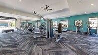 Located at Crest Village in Flowery Branch, Georgia, this single-floor fitness facility offers a generously sized, thoughtfully designed space for comprehensive wellness and exercise. The exercise room is the centerpiece of the facility, featuring bold teal accent walls, herringbone-patterned carpet flooring, and elevated ceilings fitted with ceiling fans and recessed lighting that create a bright, energizing atmosphere. Large windows line the walls, flooding the interior with natural light and offering views of the surrounding community. The room is equipped with an extensive array of fitness equipment, including treadmills, ellipticals, rowing machines, cable machines, weight benches, and free weights, accommodating a full range of cardio and strength training workouts. Full-length mirrors along one wall enhance the sense of space and provide functional support for form-focused training. A dedicated storage area with built-in cubbies adds organizational convenience. A striking glass garage-style door opens directly onto a covered outdoor deck, seamlessly connecting the interior to the resort-style pool area beyond. The expansive deck features a beadboard ceiling, ceiling fans, and white painted columns, overlooking lounge seating, umbrellas, and a pergola-covered outdoor gathering space — making this fitness center a true community amenity destination.
