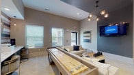 Located at 3055 Carrington Mill Blvd in Morrisville, NC, this single-floor amenity lounge is a thoughtfully designed community space that blends entertainment, comfort, and function. The open-concept layout features herringbone-patterned flooring and a neutral color palette accented by warm and dark tones that lend a contemporary, upscale atmosphere throughout.

The game area is a central highlight, equipped with a sleek pool table beneath a modern rectangular pendant light, a full-size shuffleboard table, and a large chess set on a round coffee table. Statement lighting fixtures, including sculptural chandelier-style pendants, are found throughout the space, adding visual interest to the vaulted and tray ceilings above.

A comfortable lounge seating area features a large sectional sofa adorned with decorative pillows, a wall-mounted flat-screen TV, and natural light streaming through multiple windows with views of the surrounding landscape. Adjacent to the lounge, a business center alcove offers desktop computer workstations set against a striking striped accent wall with a round mirror, providing a focused workspace within the communal setting.

High-top bar seating and a white brick accent column further define the entertainment zones, while curated wall art and mirrors add a polished, residential feel. The space is ideal for socializing, recreation, and productivity alike.