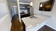 This single-floor space showcases a well-appointed kitchen as its centerpiece, designed with both functionality and aesthetics in mind. The kitchen features granite countertops with a speckled pattern in neutral tones, providing ample prep and workspace. A large peninsula with an integrated stainless steel sink serves as a natural divider, enhancing the open-concept layout while offering additional seating or workspace potential. Pendant lighting hangs above the peninsula, adding focused illumination and a polished design touch.

Dark espresso cabinetry lines the kitchen walls, offering generous storage space and creating a striking contrast against the lighter countertops. Stainless steel appliances, including a refrigerator, gas range, and built-in microwave, complete the kitchen's modern aesthetic. Rich, dark hardwood flooring extends throughout the visible areas of the space, adding warmth and continuity. A columned partition subtly defines the kitchen zone without fully enclosing it. A hallway visible in the background suggests additional rooms beyond, hinting at a well-organized and spacious overall floor plan.