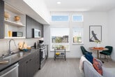 This thoughtfully designed single-floor residence offers a cohesive blend of modern aesthetics and practical functionality throughout its well-organized layout. The open-concept living area serves as the heart of the home, featuring light wood-tone flooring, clean white walls, and a wall-mounted split HVAC unit. The space comfortably accommodates a seating arrangement centered around a media console, making it an inviting area for relaxing or entertaining.

The kitchen is outfitted with dark wood-grain cabinetry, sleek gray countertops, and stainless steel appliances including a range, over-the-range microwave, and dishwasher. Open upper shelving provides both display and storage opportunities, while a single undermount sink with a modern faucet anchors the clean, linear design.

The bedroom offers a cozy retreat with a textured accent wall, soft lighting, and ample space for a full-sized bed. A sliding barn-style door adds a contemporary architectural touch to the hallway connecting the main living areas.

The bathroom features a wide vanity with gray stone-look countertops, an undermount sink, and a large mirror. A glass-enclosed shower stall completes the space. Conveniently located adjacent to the bathroom, a stacked washer and dryer unit provides full in-unit laundry capabilities, adding a highly practical element to the residence.