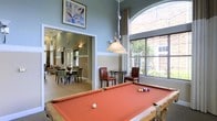 This inviting communal space is designed for relaxation and social interaction, offering a thoughtfully arranged layout across a single floor. The centerpiece of the main recreation area is a full-sized billiards table, positioned beneath an elegant pendant light that draws the eye upward toward impressively tall ceilings. The walls are finished in a calming blue-gray tone, complemented by crisp white chair-rail molding that adds a classic architectural detail throughout the space. A large arched window floods the room with natural light, offering views of lush greenery outside, while additional windows fitted with wood blinds and draped curtains contribute to the bright, airy atmosphere. Casual seating with leather chairs and bistro-style tables is thoughtfully placed to encourage conversation and leisure. Through an open doorway, an adjacent room reveals a dining area with multiple tables and chairs, ideal for gatherings and social events. Wall sconces and framed artwork add warmth and character to the overall design. The open, flowing layout and generous ceiling height create a sense of spaciousness, making this a versatile and comfortable environment for residents or guests to enjoy.