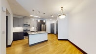 This single-floor residence is designed with a clean, contemporary aesthetic that balances functionality and style. The open-concept kitchen and living area serves as the heart of the unit, featuring warm light hardwood flooring that extends throughout. The kitchen is well-appointed with a two-toned cabinet scheme — light gray upper cabinets paired with darker lower cabinetry — complemented by a white tile backsplash and a full suite of stainless steel appliances, including a French-door refrigerator, electric range, and built-in microwave. A central island with a granite countertop and undermount sink provides additional prep space and casual seating. Pendant lighting suspended above the island adds a stylish focal point, while a statement pendant fixture near the entryway enhances the welcoming atmosphere.

The living area benefits from natural light through large windows and a glass door leading to an outdoor access point, with a ceiling fan overhead for added comfort. Bedrooms feature plush neutral carpeting, ceiling fans, and windows that bring in natural light. The bathroom includes a soaking tub framed by marble-style tile surround, hardwood-style flooring, and gray cabinetry for storage. Throughout the space, dark baseboards provide a sharp contrast against bright white walls, contributing to the unit's modern, polished character.