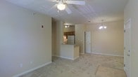This single-floor residence offers a comfortable and functional layout centered around an open-concept living space. The main living area is generously sized, featuring plush wall-to-wall carpeting in a neutral tone that adds warmth underfoot. A ceiling fan with a light fixture sits prominently overhead, providing both airflow and ambient lighting, while a decorative chandelier adds an elegant touch to the dining area nearby.

The kitchen is positioned at the heart of the home, separated from the living space by a raised breakfast bar that creates a natural division while maintaining an open feel. The kitchen is equipped with wood-toned cabinetry, a refrigerator, and a range, with track lighting above illuminating the workspace effectively. The pass-through design ensures the kitchen remains connected to the main living area, making the space ideal for everyday living and casual entertaining.

A doorway visible from the main living area leads to additional rooms, suggesting a well-organized floor plan. The walls are finished in a soft, neutral beige tone throughout, complementing the light-colored carpeting and creating a cohesive, understated aesthetic. Clean white baseboards and trim provide a crisp contrast, adding a polished finish to the overall interior.