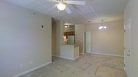 This single-floor residence offers a comfortable and functional layout centered around an open-concept living space. The main living area is generously sized, featuring plush wall-to-wall carpeting in a neutral tone that adds warmth underfoot. A ceiling fan with a light fixture sits prominently overhead, providing both airflow and ambient lighting, while a decorative chandelier adds an elegant touch to the dining area nearby.

The kitchen is positioned at the heart of the home, separated from the living space by a raised breakfast bar that creates a natural division while maintaining an open feel. The kitchen is equipped with wood-toned cabinetry, a refrigerator, and a range, with track lighting above illuminating the workspace effectively. The pass-through design ensures the kitchen remains connected to the main living area, making the space ideal for everyday living and casual entertaining.

A doorway visible from the main living area leads to additional rooms, suggesting a well-organized floor plan. The walls are finished in a soft, neutral beige tone throughout, complementing the light-colored carpeting and creating a cohesive, understated aesthetic. Clean white baseboards and trim provide a crisp contrast, adding a polished finish to the overall interior.