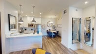 This single-floor residence showcases a thoughtfully designed open-concept layout that seamlessly connects the kitchen, dining, and living areas. The kitchen is a standout feature, equipped with crisp white shaker cabinetry extending to the ceiling, stainless steel appliances including a range, built-in microwave, and refrigerator, a white subway tile backsplash, and a generously sized central island topped with a marble-look quartz countertop. Dual industrial-style pendant lights hang above the island, adding a modern touch. The dining area is defined by a round wooden table surrounded by contemporary chairs, accented by decorative wall clocks and warm pendant lighting. The living area features hardwood-style flooring that flows throughout the main living spaces, with a wall-mounted television above a stylish media console. The bedroom offers plush carpeting, ample natural light through shuttered windows, a ceiling fan, and two sleek dark-paneled doors providing access to closet and bathroom spaces. The bathroom is finished with large-format tile flooring, a floating vanity with quartz countertops, and a full bathtub with shower. Clean white walls and recessed lighting throughout create a bright, welcoming ambiance.